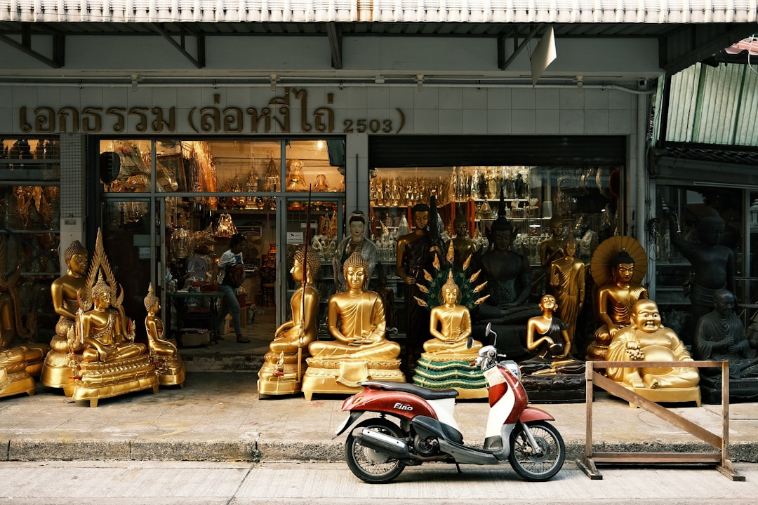 Thai Buddhist statue shop storefront with golden Buddha figures and a red scooter parked outside
