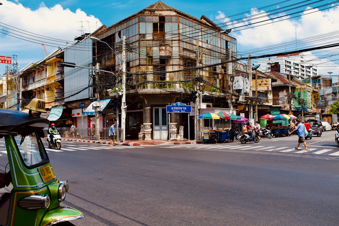 Historic street corner in Thailand with weathered colonial building, green tuk-tuks, and street vendors under colorful umbrellas